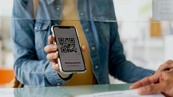 Person showing a smartphone with a QR code and a digital queue ticket display at a counter, symbolizing digital services in the public sector.