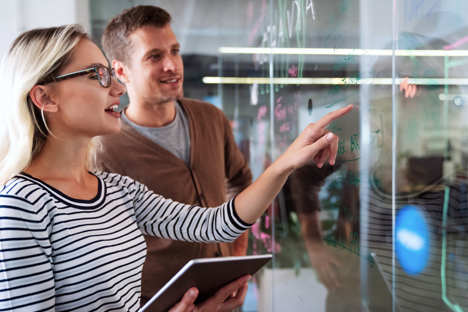 Planning Cloud Migration Two people standing in front of a glass wall discussing a plan. One person points to notes and diagrams written on the wall.