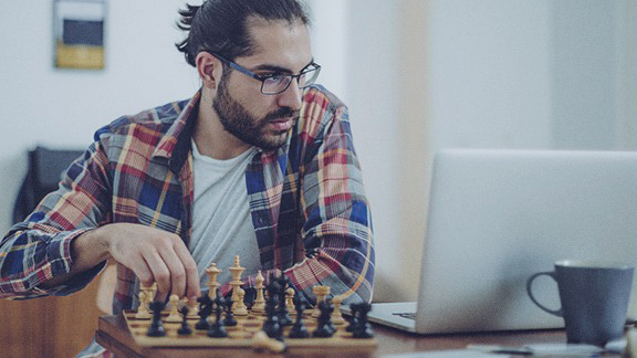 A young man is sitting at home in front of his laptop with a chessboard next to him. He is learning online chess.
