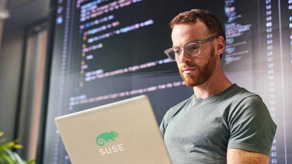 IT-Experte arbeitet an einem Laptop vor Code-Anzeigen auf Bildschirmen. Das Bild steht für ein modernes Cloud-Betriebssystem-Upgrade mit SUSE Linux.