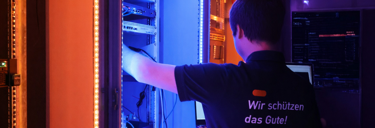 IT professional working on an open server rack in a server room illuminated with blue and orange light. The shirt reads “Wir schützen das Gute!”.
