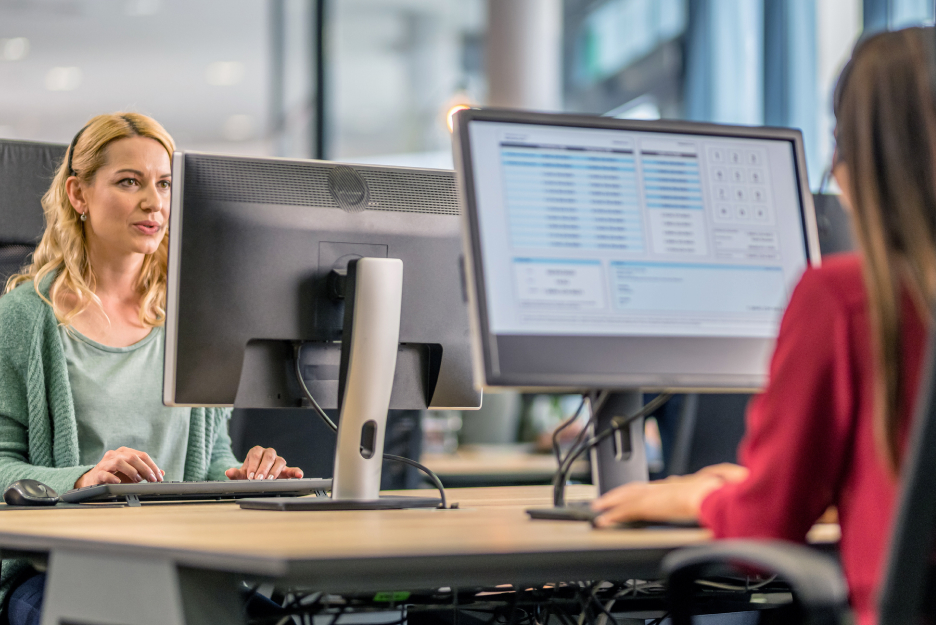 Zwei Personen arbeiten an Computerarbeitsplätzen in einem Büro. Im Vordergrund sind Monitore mit geöffneten Anwendungen zu sehen, während eine Person auf einen Bildschirm blickt.
