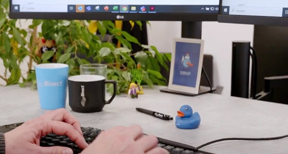 Person tippt auf Tastatur an einem Büroarbeitsplatz mit Binect-Tassen und zwei Monitoren im Hintergrund