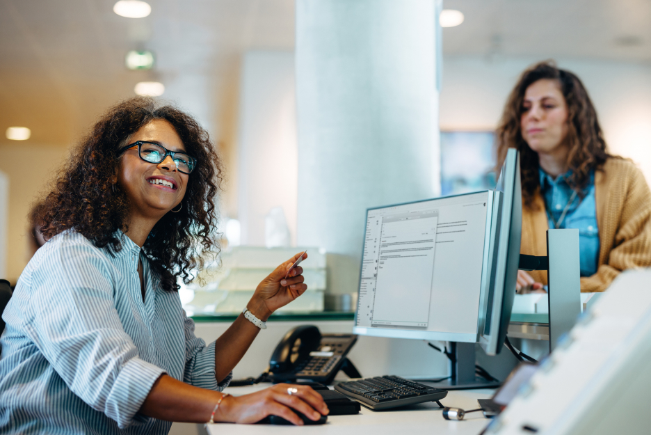 Mitarbeiterin arbeitet an einem Computerarbeitsplatz in einer modernen Büroumgebung, während eine weitere Person im Hintergrund an einem Verwaltungsbereich steht.