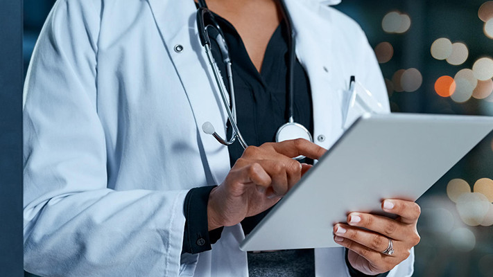 A doctor in a white coat uses a tablet, with blurred lights in the background.