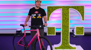 Ein Mann hält ein Fahrrad neben einem mit Moos bewachsenem großen Telekom Logo.