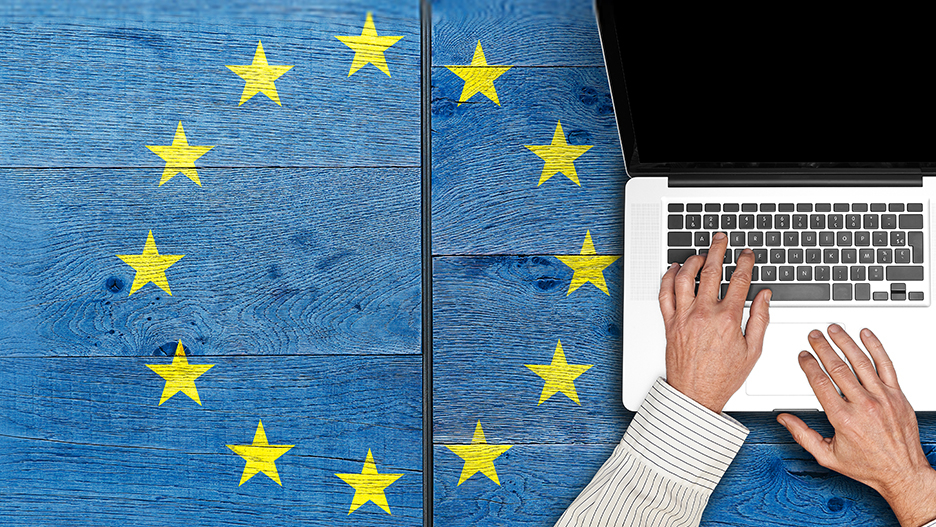 Person tippt auf einer Laptop-Tastatur auf einem blauen Holztisch, umgeben von gelben Sternen der EU Flagge