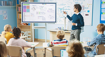 Schüler sitzen in einem Klassenraum, ein Lehrer steht vor einer digitalen Tafel 